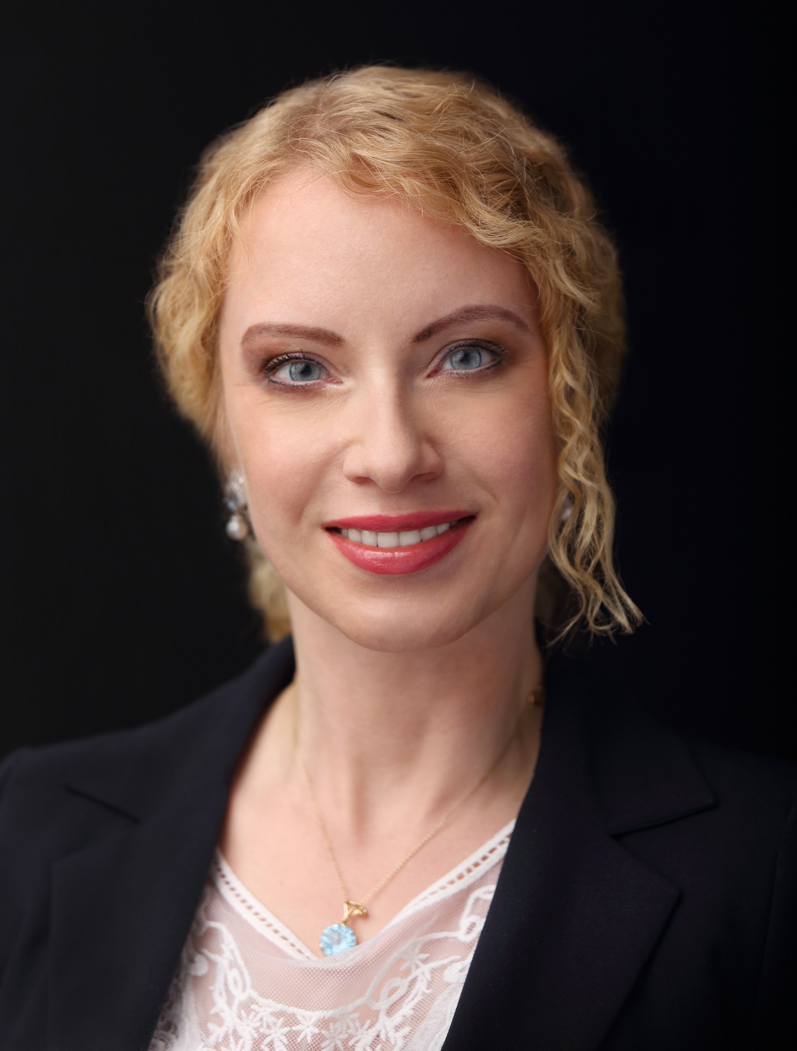 Portret uśmiechniętej kobiety w czarnej marynarce na ciemnym tle. Profesjonalny wizerunek adwokata z Warszawy, z blond włosami i delikatną biżuterią.