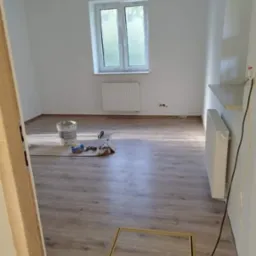 Pokój po remoncie z drewnopodobną podłogą, białymi ścianami, grzejnikiem pod oknem i puszką farby na podłodze. Widoczna rama drzwiowa bez drzwi oraz niezamontowana półka.