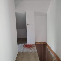 aleksander kowalski - Poddasze po remoncie, świeżo pomalowane na biało, z widocznym fragmentem drewnianej konstrukcji podłogi i zwiniętym pomarańczowym kablem na podłodze imitującej drewno.