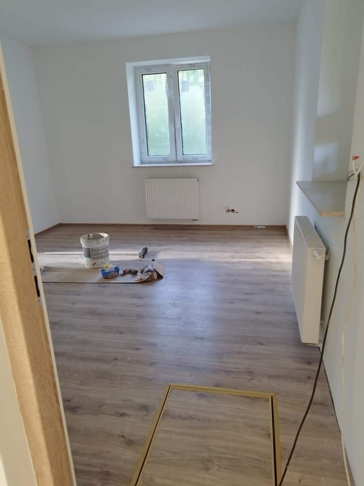 Pokój po remoncie z drewnopodobną podłogą, białymi ścianami, grzejnikiem pod oknem i puszką farby na podłodze. Widoczna rama drzwiowa bez drzwi oraz niezamontowana półka.