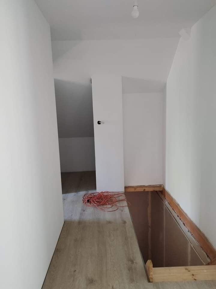 Poddasze po remoncie, świeżo pomalowane na biało, z widocznym fragmentem drewnianej konstrukcji podłogi i zwiniętym pomarańczowym kablem na podłodze imitującej drewno.