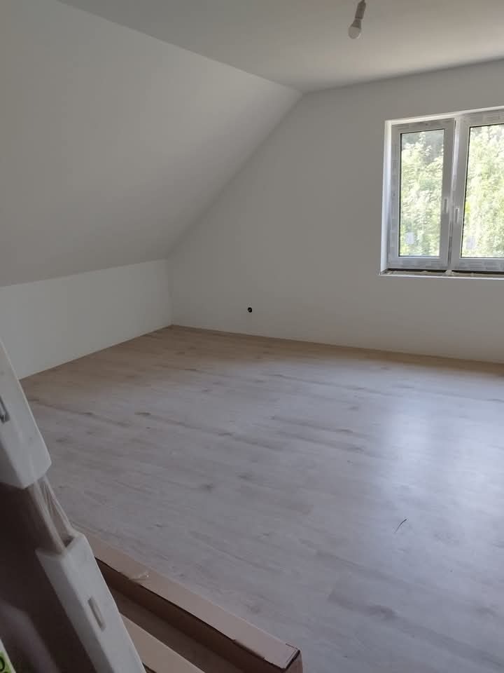 Poddasze po remoncie z nową podłogą laminowaną o jasnym odcieniu drewna, widoczne okno i białe ściany, częściowo zasłonięte opakowania po panelach podłogowych w rogu kadru.