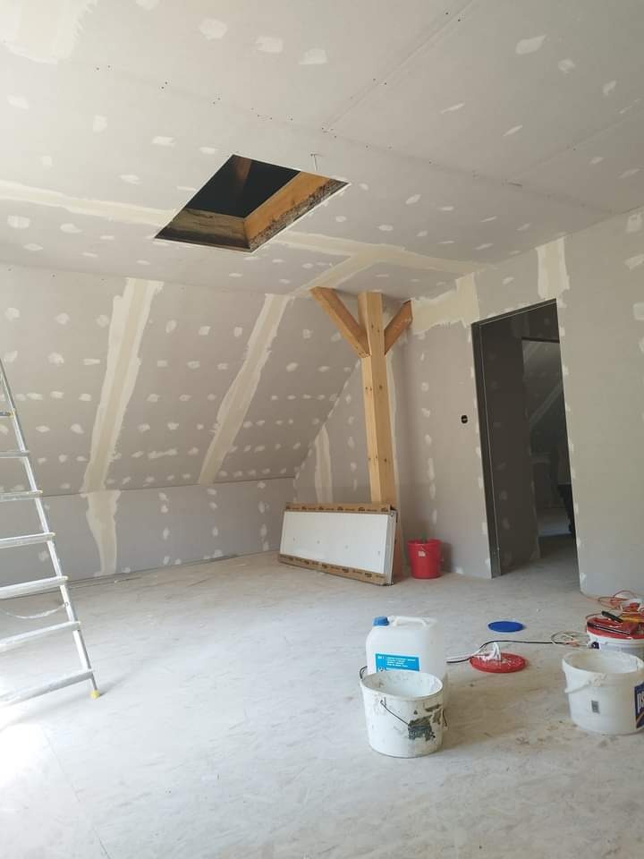 Poddasze w trakcie prac wykończeniowych: ściany pokryte płytami gipsowo-kartonowymi z widocznymi łączeniami wymagającymi szpachlowania, otwór okienny w suficie, drewniany element konstrukcyjny...