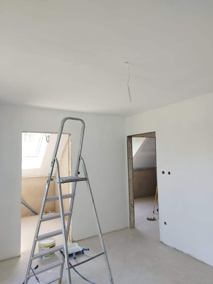 Wnętrze po szpachlowaniu ścian i sufitu, widoczna aluminiowa drabina, wiadro z farbą i niedokończone otwory na gniazdka elektryczne.
