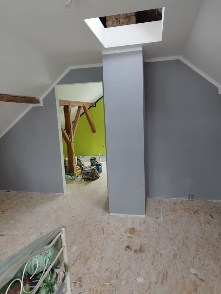 Pomalowane na szaro ściany poddasza z drewnianą konstrukcją dachu widoczną w otworze drzwiowym, surowa podłoga z płyty OSB i okno dachowe w suficie.