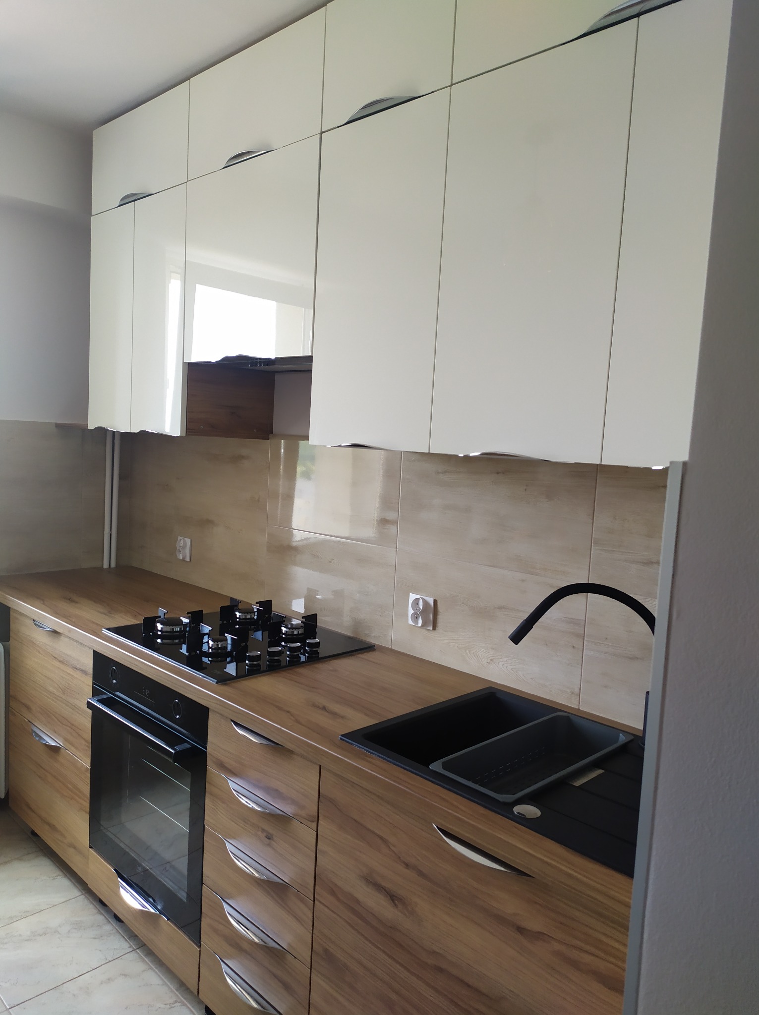 Nowoczesna kuchnia z białymi szafkami górnymi i drewnopodobnymi szafkami dolnymi, czarny zlew i płyta gazowa, jasne płytki na ścianie.