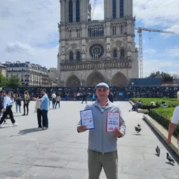 Mężczyzna w dżokejce prezentuje przewodniki po Francji i Rata Touille na tle katedry Notre-Dame w Paryżu, z widocznymi rusztowaniami i turystami w tle.
