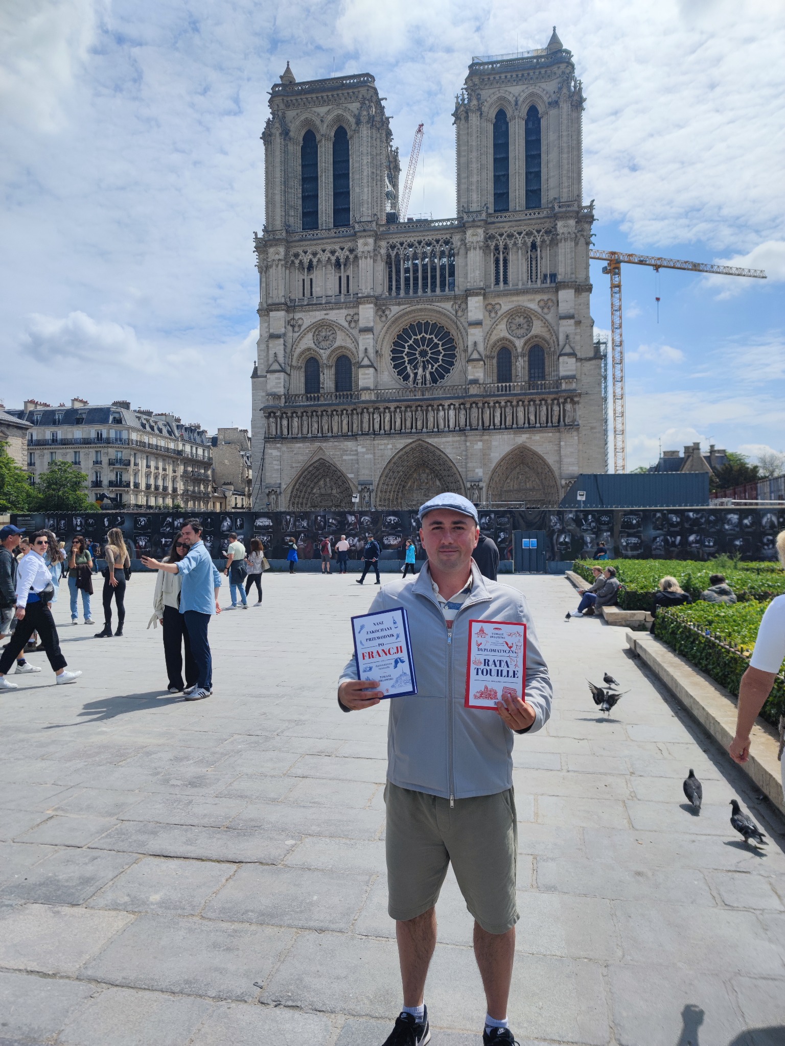 Mężczyzna w dżokejce prezentuje przewodniki po Francji i Rata Touille na tle katedry Notre-Dame w Paryżu, z widocznymi rusztowaniami i turystami w tle.