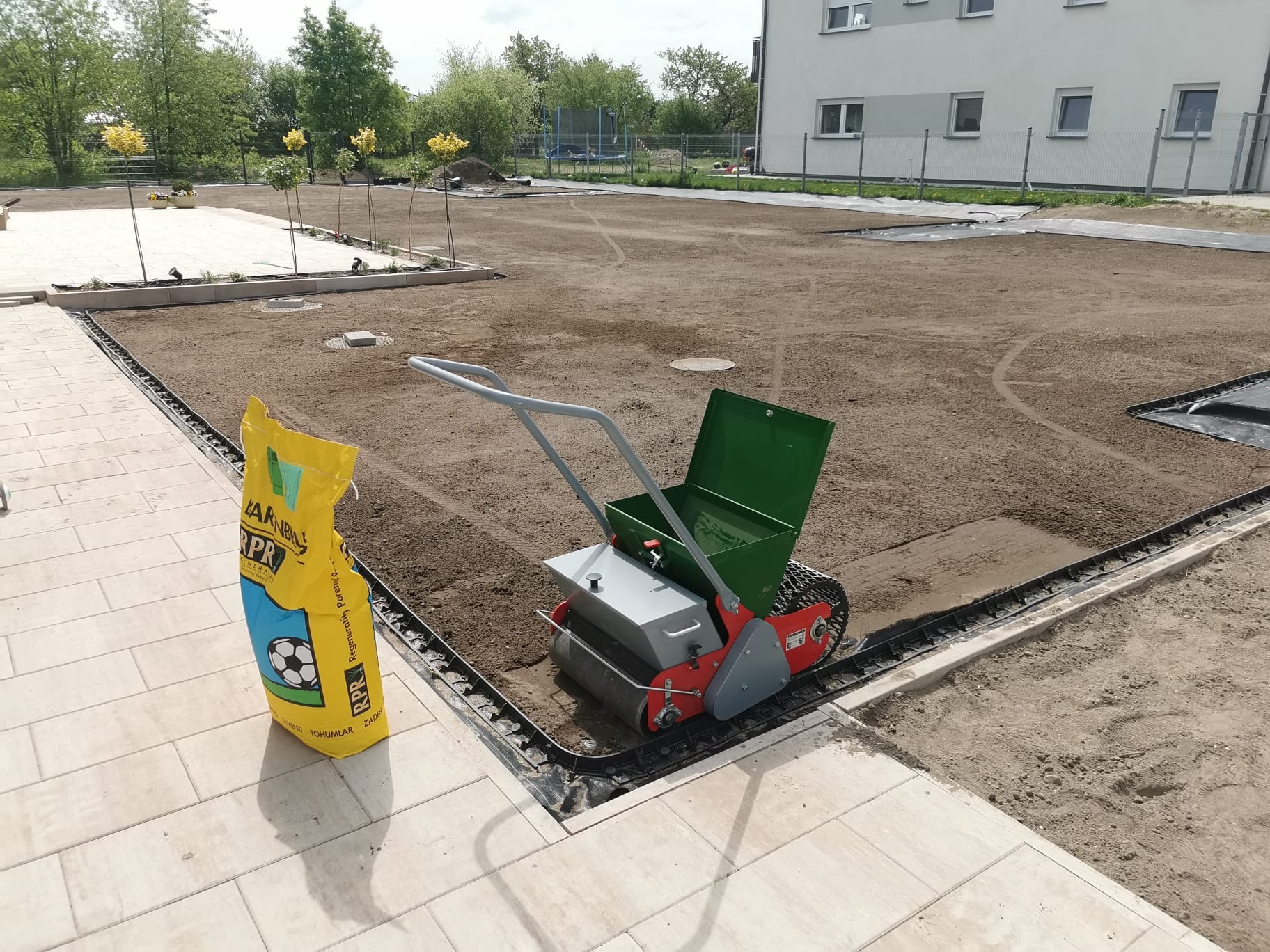 Przygotowanie terenu pod trawnik: siewnik do trawy i worek z nasionami na tle wyrównanej ziemi i nowej kostki brukowej. Widoczne drzewka i budynek w tle.