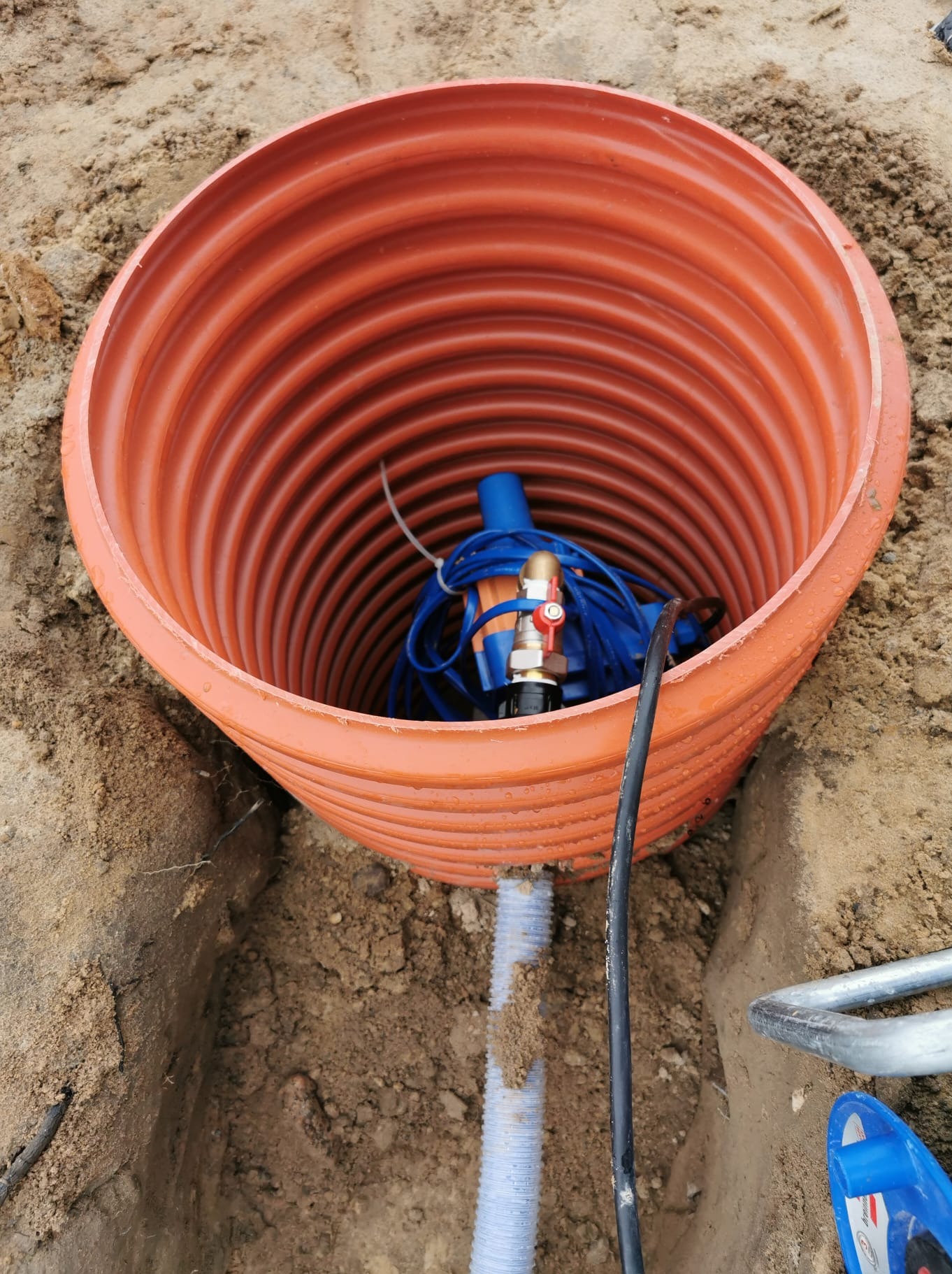 Pomarańczowa rura karbowana w wykopie, wewnątrz niebieski system nawadniania z przewodami. Montaż instalacji nawadniającej w ziemi.