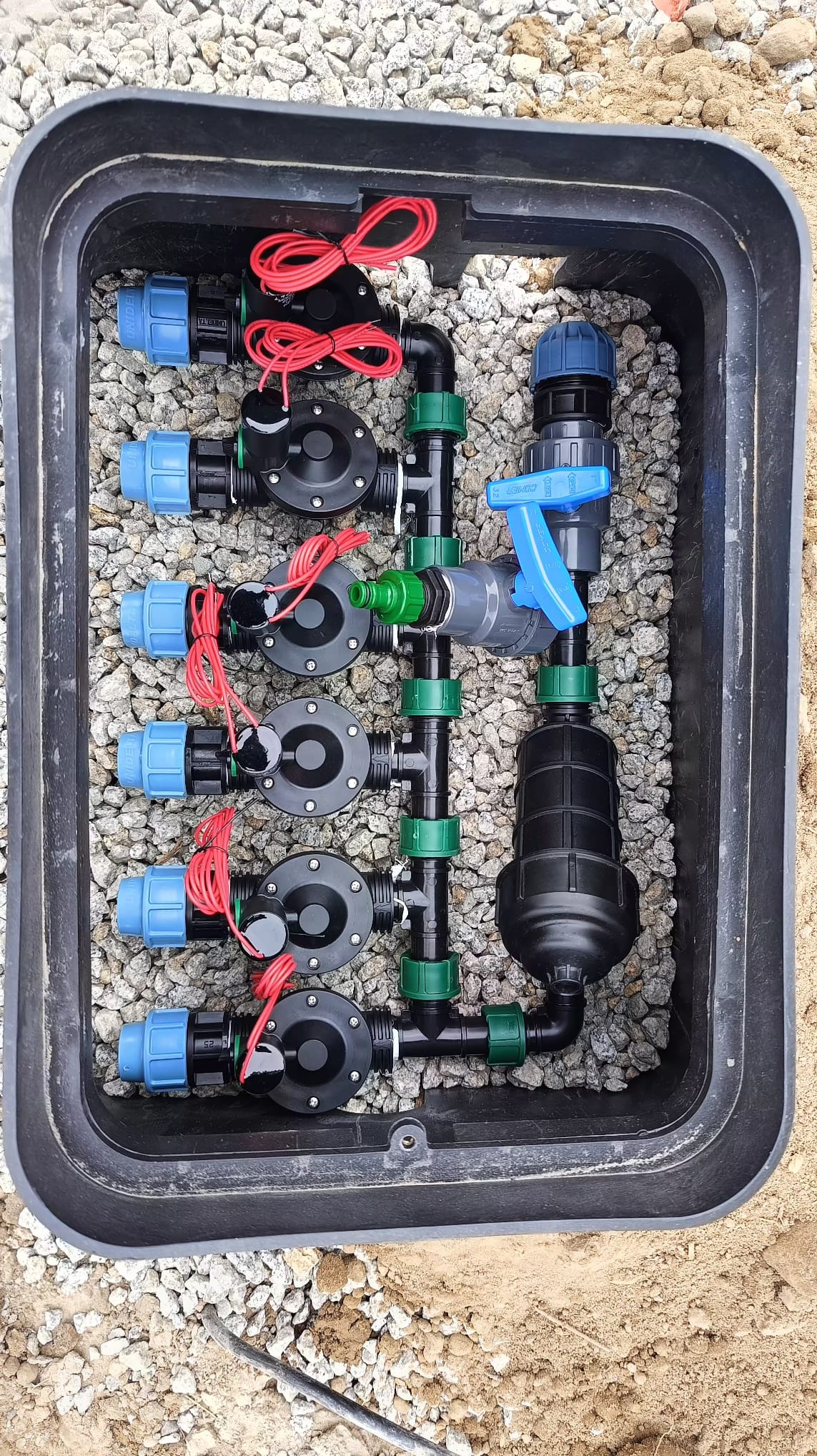 Skrzynka z zaworami i filtrem systemu nawadniania, wypełniona drobnymi kamykami. Widoczne czarne rury, niebieskie złączki i czerwone przewody.