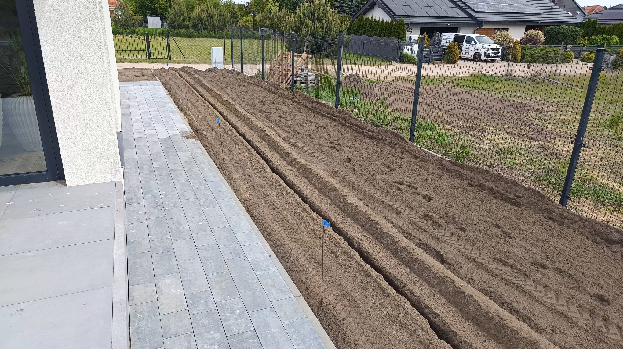 Przygotowane rowy w ziemi, obok kostki brukowej, prawdopodobnie pod system nawadniania. Widoczne ślady opon i wbite znaczniki.