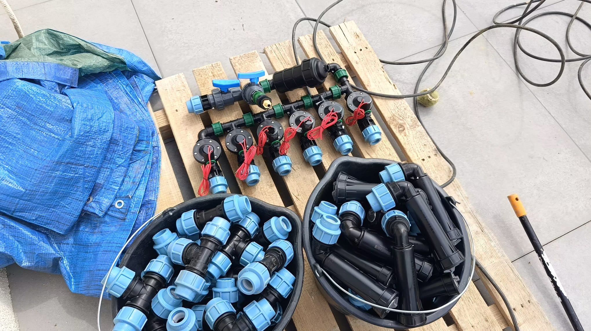 Elementy systemu nawadniania: złączki w wiadrach, rury i filtry na palecie. Przygotowanie do instalacji, widoczny kabel i plandeka.