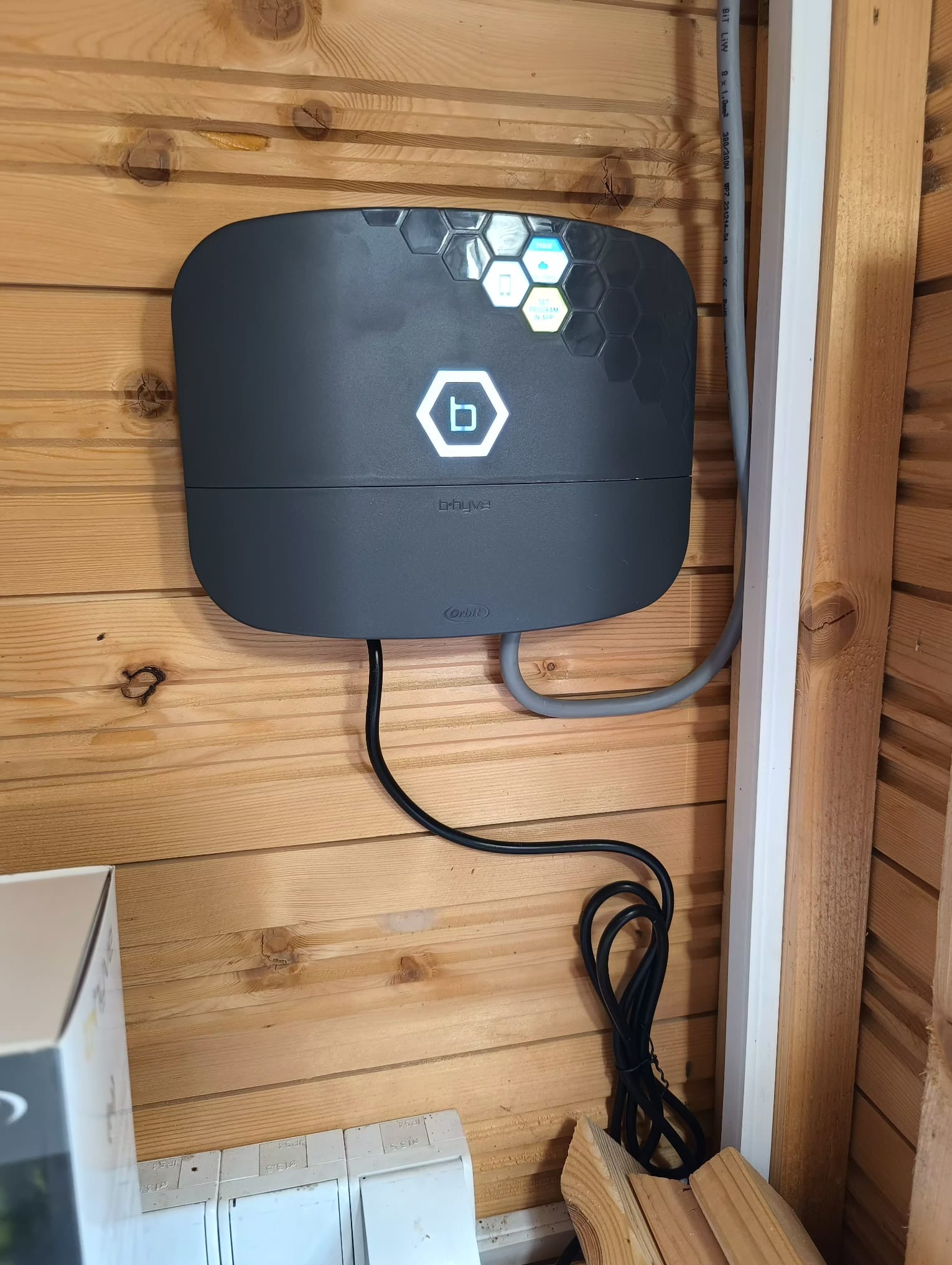 Centrala sterująca systemem nawadniania Orbit B-hyve na drewnianej ścianie. Widoczne przewody i elementy instalacji elektrycznej.