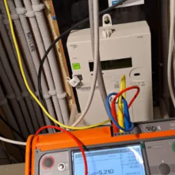Elektryk wykonuje pomiary impedancji pętli zwarcia ZL-PE=5,21Ω za pomocą miernika Sonel MPI-520, widoczny licznik energii elektrycznej Landis+Gyr E360 oraz instalacja elektryczna w peszlach.
