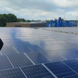 DN-ELEKTRO Daniel Niziński - Mężczyzna w bluzie z logo firmy stoi na dachu pokrytym panelami słonecznymi, obserwując panoramę miasta w oddali.