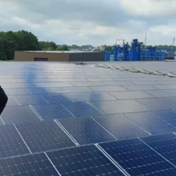 Mężczyzna w bluzie z logo firmy stoi na dachu pokrytym panelami słonecznymi, obserwując panoramę miasta w oddali.