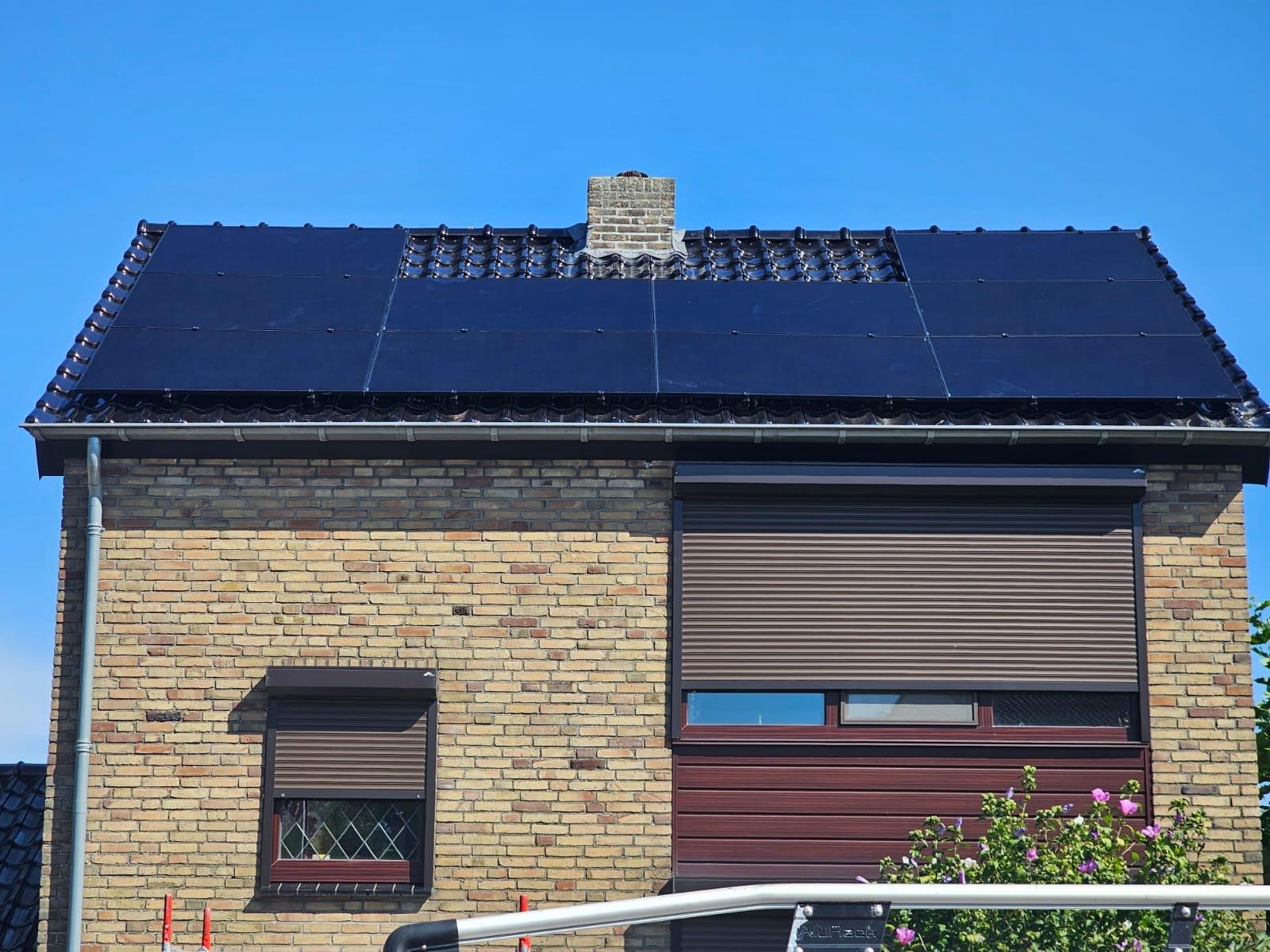Dom z cegły z ciemnymi panelami fotowoltaicznymi na dachu, widoczne okna z roletami zewnętrznymi i komin.