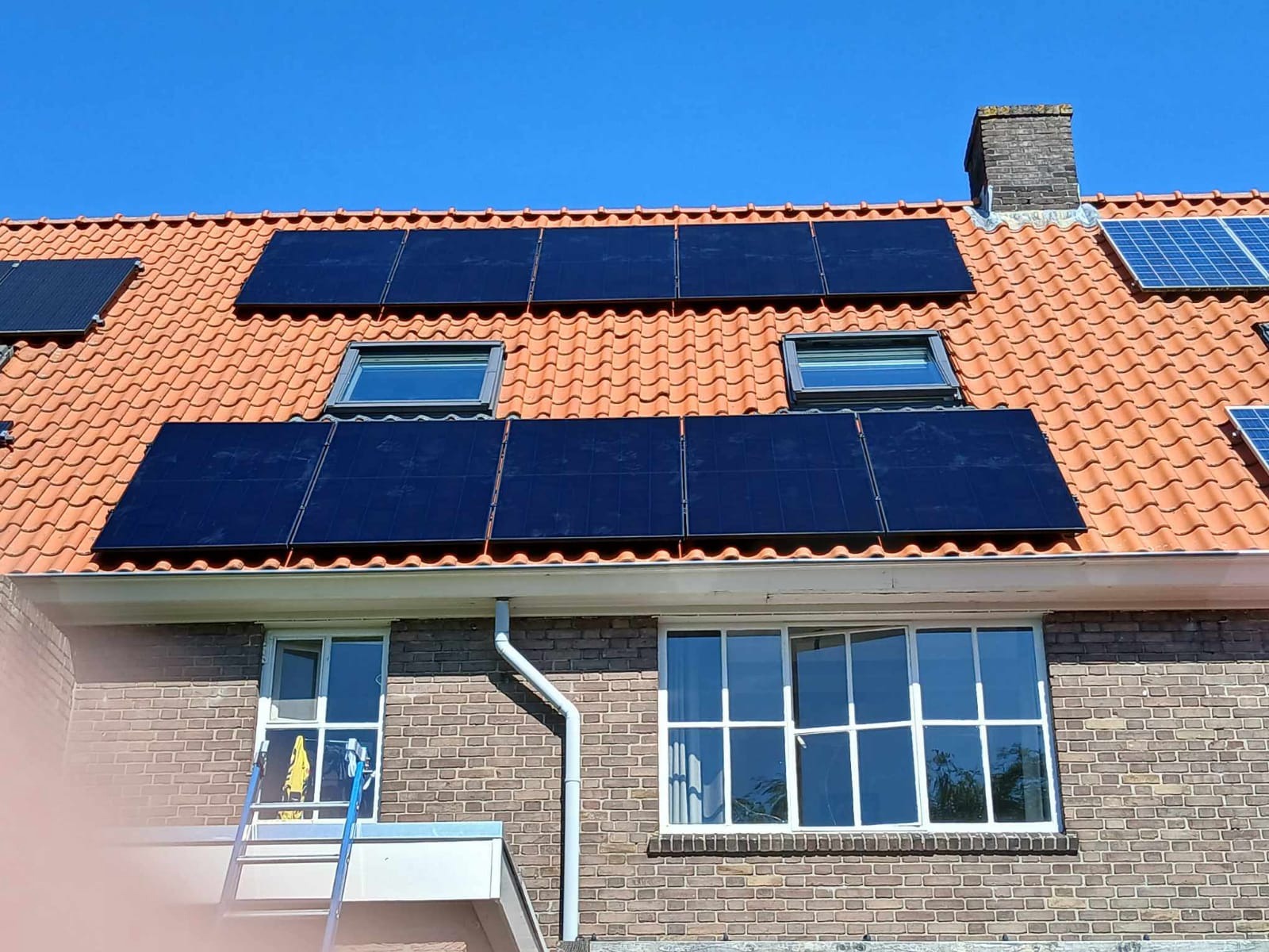 Czerwony dach z dachówki ceramicznej z zamontowanymi panelami fotowoltaicznymi i oknami dachowymi, widoczne okno budynku z drabinką opartą o ścianę.