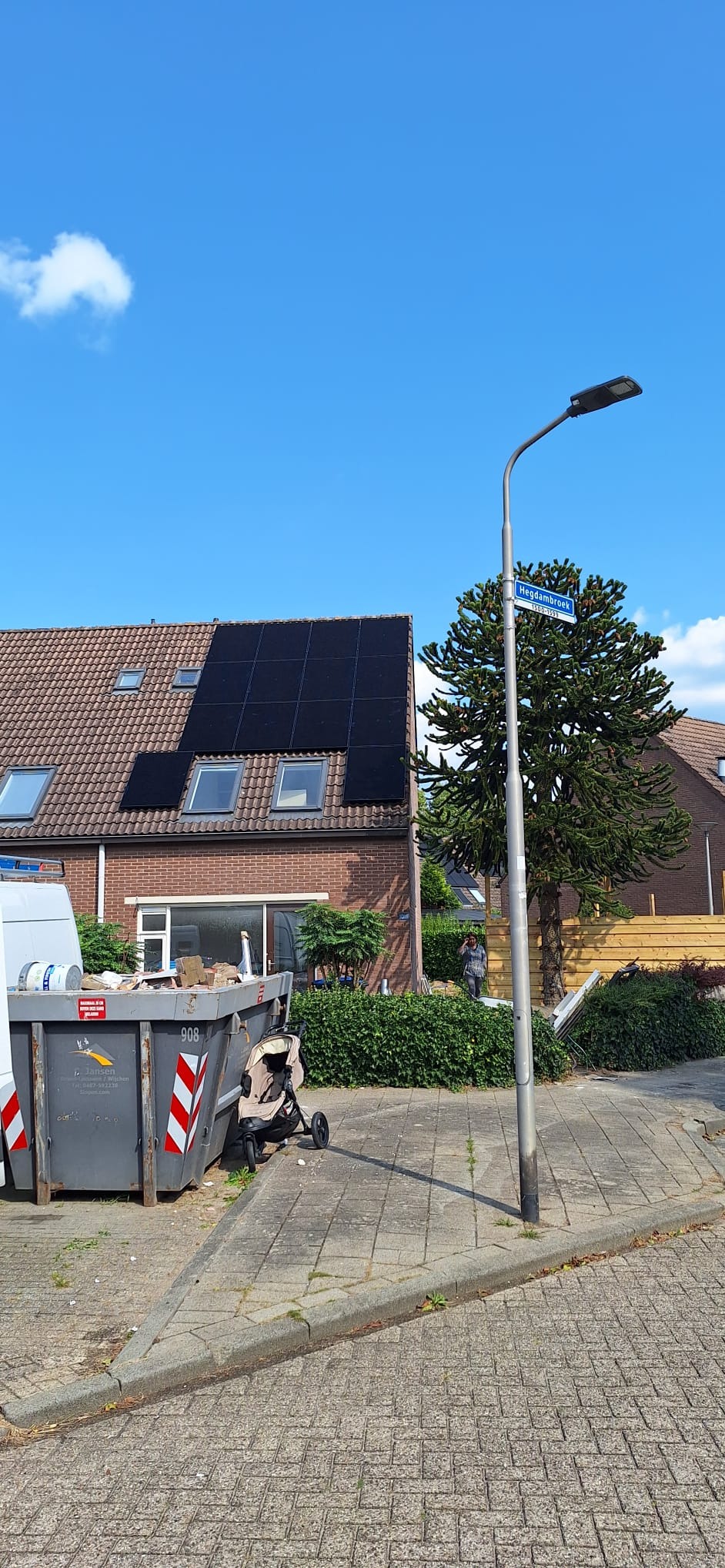 Dom z panelami słonecznymi na dachu, widoczny kontener na śmieci, w tle osoba przy ogrodzeniu i tablica z nazwą ulicy Hegdambroek.