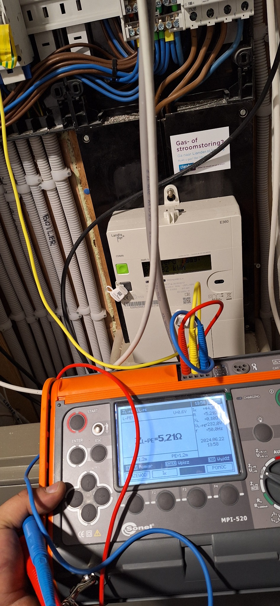 Elektryk wykonuje pomiary impedancji pętli zwarcia ZL-PE=5,21Ω za pomocą miernika Sonel MPI-520, widoczny licznik energii elektrycznej Landis+Gyr E360 oraz instalacja elektryczna w peszlach.