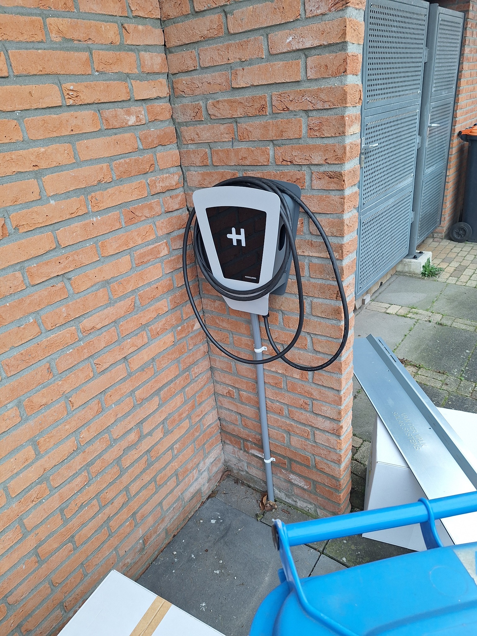 Szaro-biała stacja ładowania samochodów elektrycznych zamontowana na ścianie z czerwonej cegły, z widocznym kablem i literą 'H' na froncie, na zewnątrz budynku.
