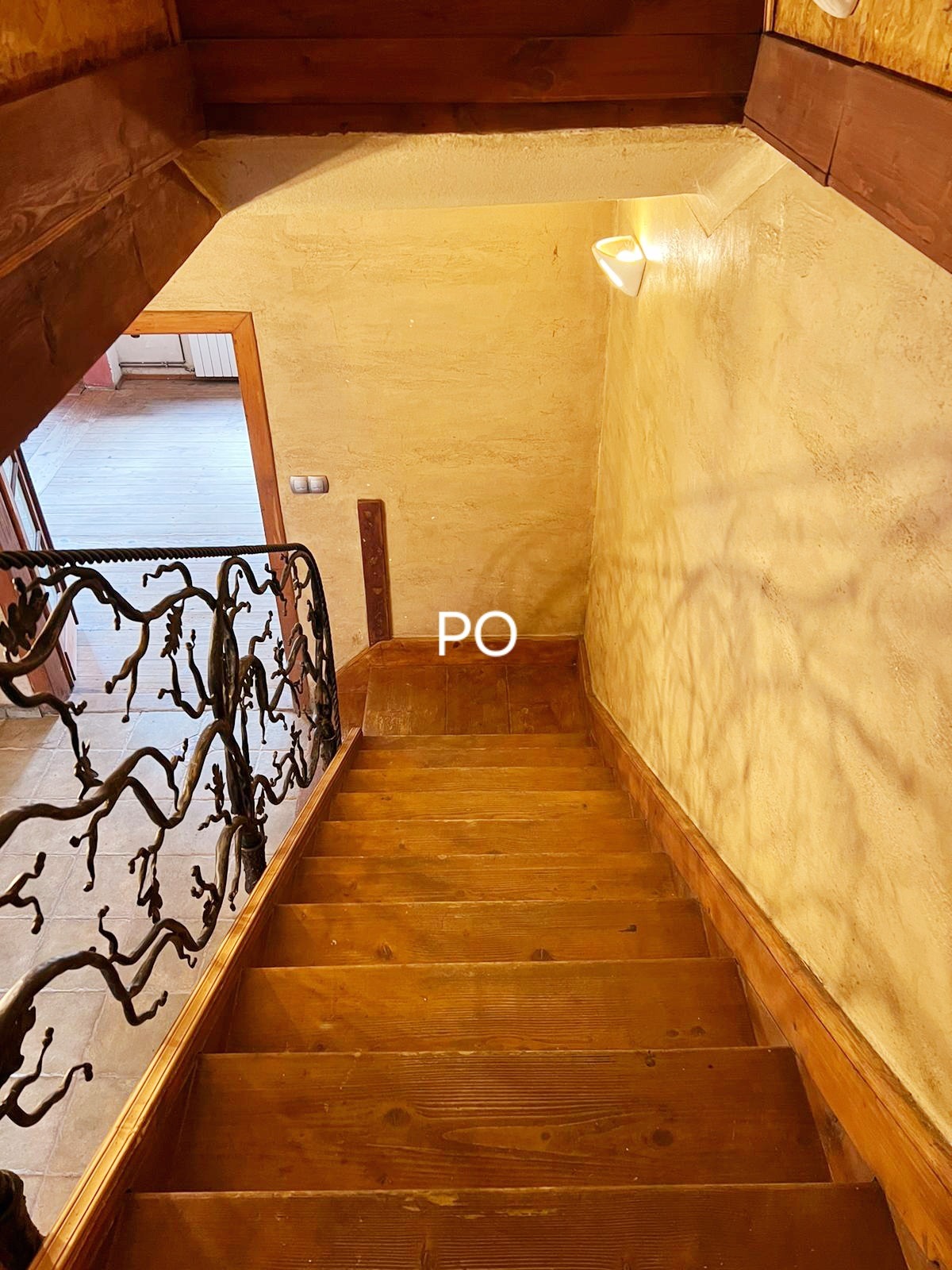 Drewniane schody z ozdobną, metalową balustradą w kształcie gałęzi drzew, prowadzące na górę, widoczne oświetlenie ścienne i fragment podłogi na górze.