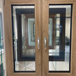 Fasada Windows & Doors - Dwuskrzydłowe okno z drewnopodobną ramą, widoczne uchwyty i ekspozycja innych modeli w tle.