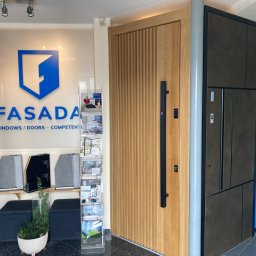 Fasada Windows & Doors - Ekspozycja dwóch modeli drzwi w salonie: jasne drzwi z pionowymi frezami i długim czarnym uchwytem oraz ciemne drzwi z panelem kolorów Gealan.