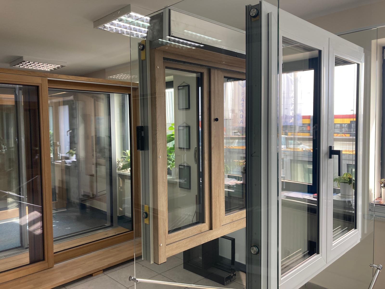 Ekspozycja w salonie: przesuwne okno drewniane z czarną ramą, okno PCV z białymi ramami i standardowe okno drewniane z widocznymi szybami i roślinami w tle.