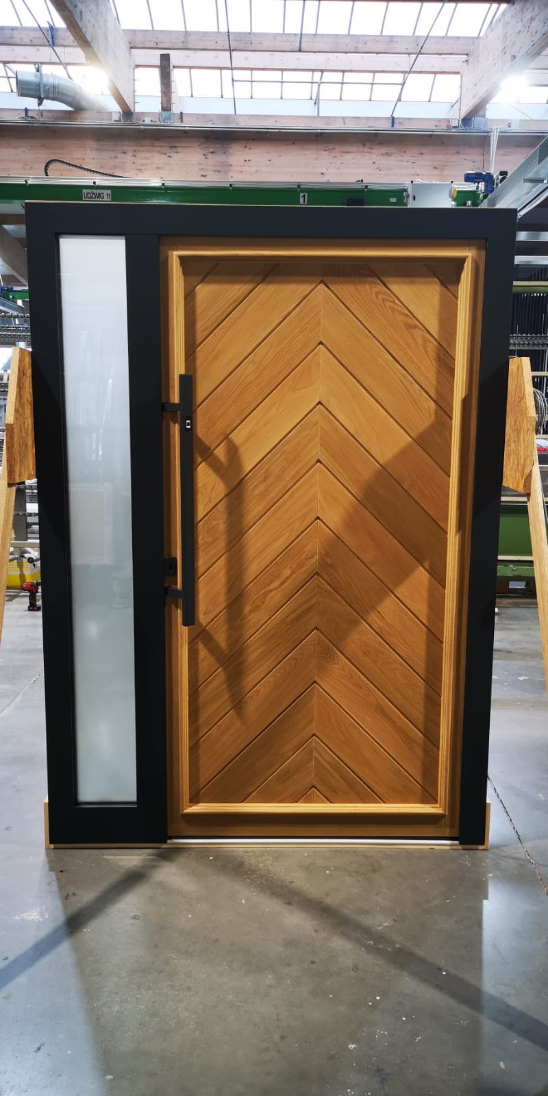 Nowoczesne drzwi przesuwne z drewna z wzorem chevron, czarna rama i szklany panel boczny, prezentowane w hali produkcyjnej.