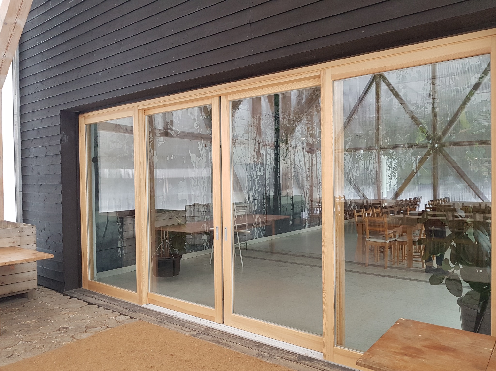 Zestaw trzech drewnianych drzwi przesuwnych z widokiem na przestrzeń restauracyjną z drewnianymi stołami i krzesłami, czarna drewniana elewacja budynku, dywanik przed wejściem.