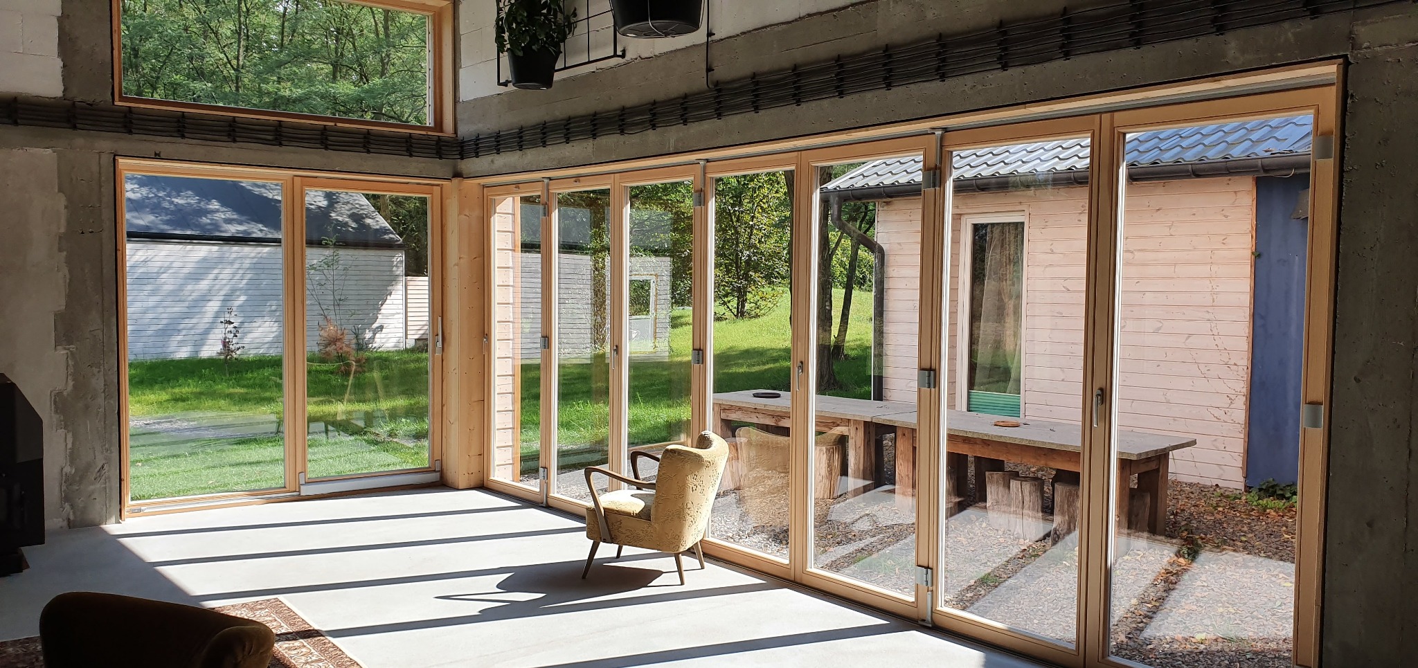 Jasne wnętrze z dużymi przesuwnymi oknami drewnianymi, z widokiem na zielony ogród i budynki gospodarcze, oświetlone promieniami słońca padającymi na betonową podłogę.