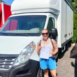 Kobieta w okularach przeciwsłonecznych trzyma dokumenty przed białym samochodem dostawczym z zabudową kontenerową w słoneczny dzień.