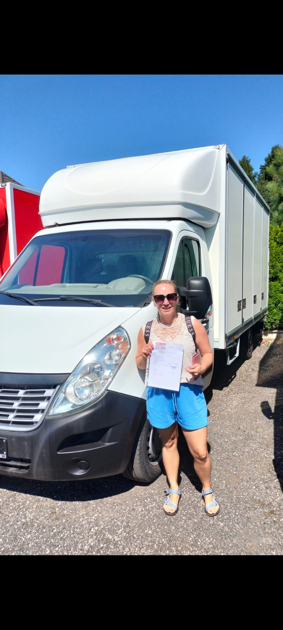Kobieta w okularach przeciwsłonecznych trzyma dokumenty przed białym samochodem dostawczym z zabudową kontenerową w słoneczny dzień.