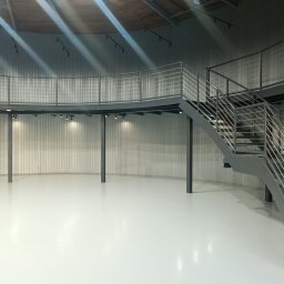 SpawajTo Grzegorz Marcinów - Wnętrze okrągłego pomieszczenia z antresolą i metalowymi schodami z balustradami ze stali nierdzewnej, oświetlone punktowymi lampami.