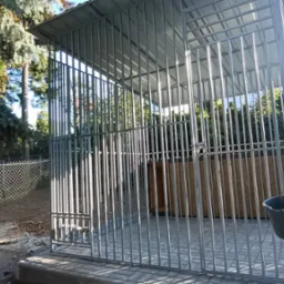 Metalowa konstrukcja klatki z zadaszeniem, z widocznym kranem i wiadrem z boku, ustawiona na kostce brukowej w ogrodzie z drzewami i siatką ogrodzeniową w tle.