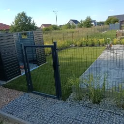 SpawajTo Grzegorz Marcinów - Nowoczesne ogrodzenie panelowe z bramką i zabudową śmietnikową w kolorze antracytowym, otoczone kamieniami ozdobnymi i zielenią, z widokiem na domy w tle.