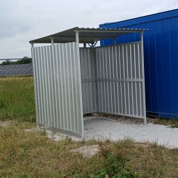 SpawajTo Grzegorz Marcinów - Metalowa konstrukcja osłonowa z zadaszeniem, wykonana z blachy trapezowej, stojąca na żwirowym podłożu obok niebieskiego kontenera, z widoczną trawą i w oddali panele słoneczne.