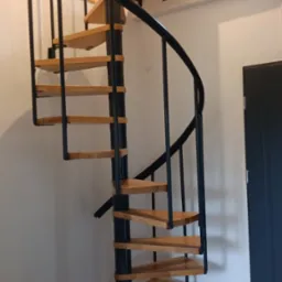Spiralne schody z drewnianymi stopniami i czarną metalową konstrukcją w jasnym wnętrzu, widoczne drzwi i drewniane żaluzje w tle.