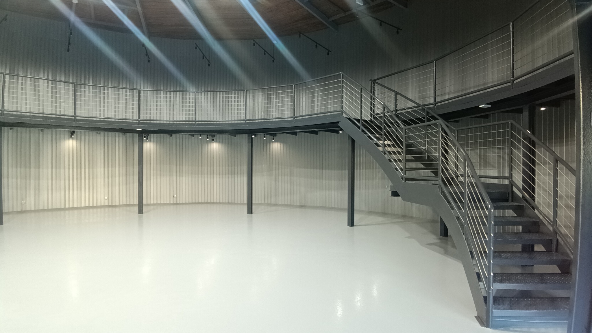 Wnętrze okrągłego pomieszczenia z antresolą i metalowymi schodami z balustradami ze stali nierdzewnej, oświetlone punktowymi lampami.