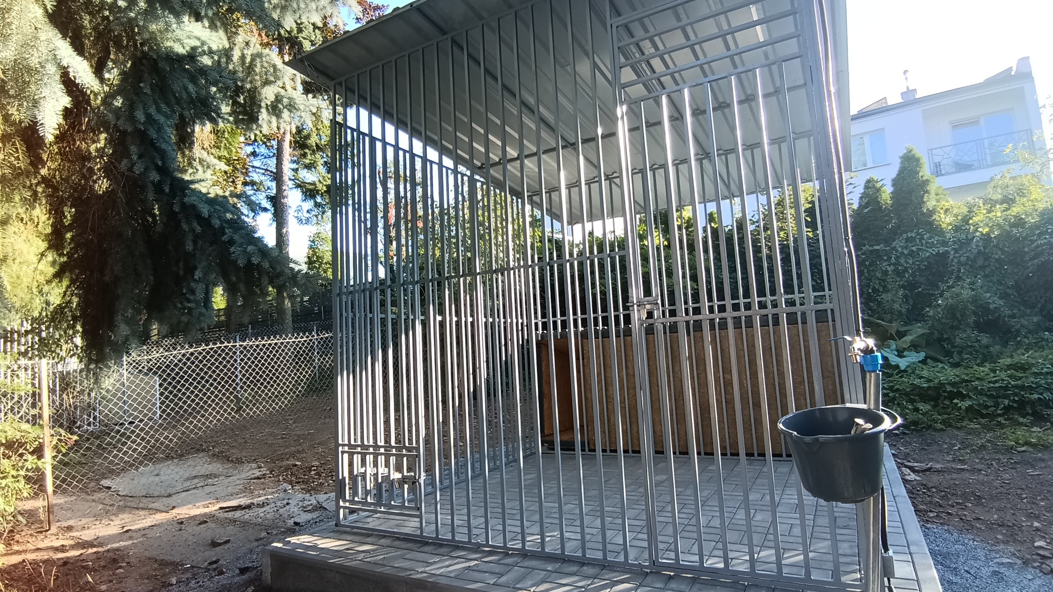 Metalowa konstrukcja klatki z zadaszeniem, z widocznym kranem i wiadrem z boku, ustawiona na kostce brukowej w ogrodzie z drzewami i siatką ogrodzeniową w tle.