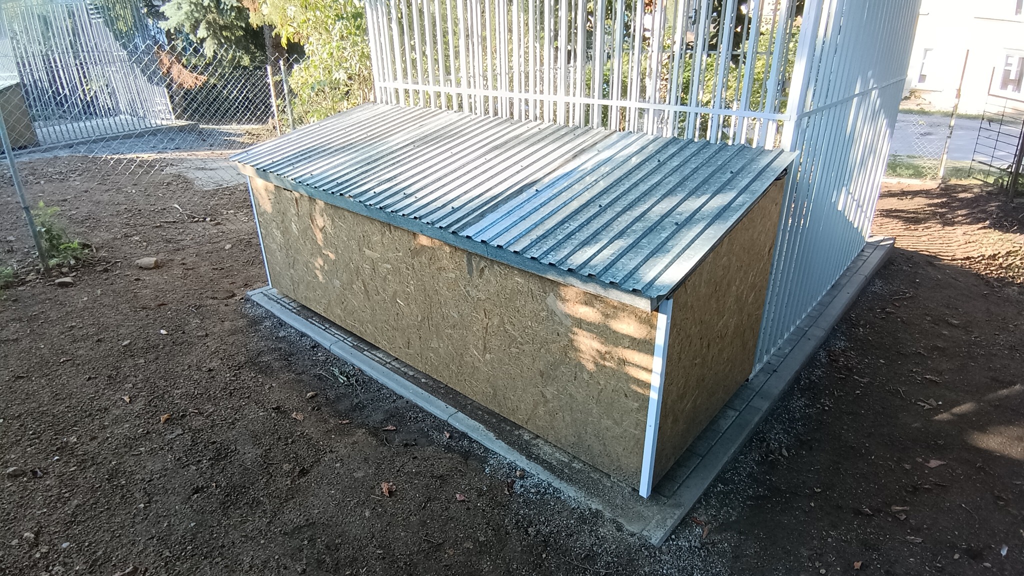 Zewnętrzna drewniana skrzynia z blachą falistą na dachu, umieszczona na betonowej podstawie przy metalowym ogrodzeniu panelowym.