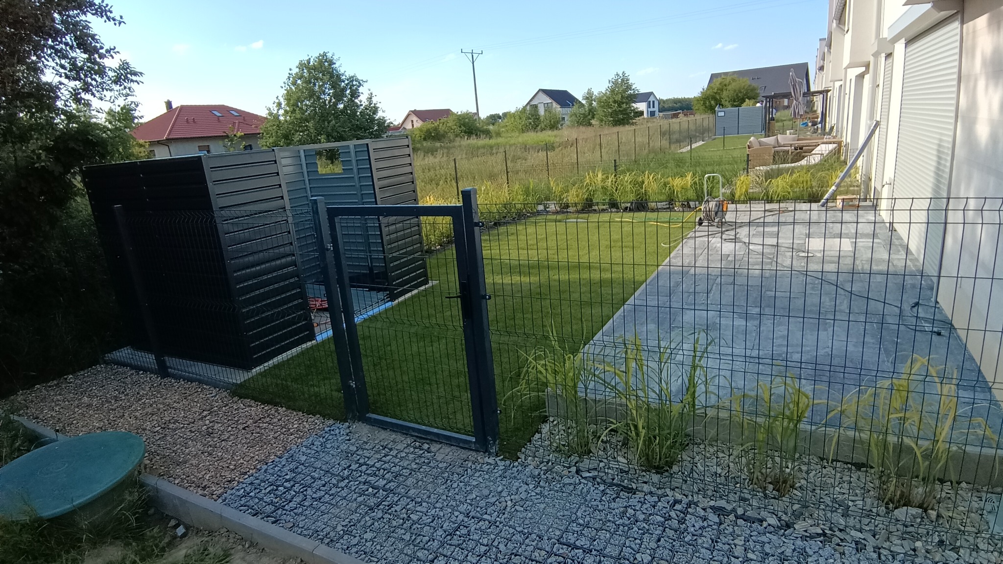 Nowoczesne ogrodzenie panelowe z bramką i zabudową śmietnikową w kolorze antracytowym, otoczone kamieniami ozdobnymi i zielenią, z widokiem na domy w tle.