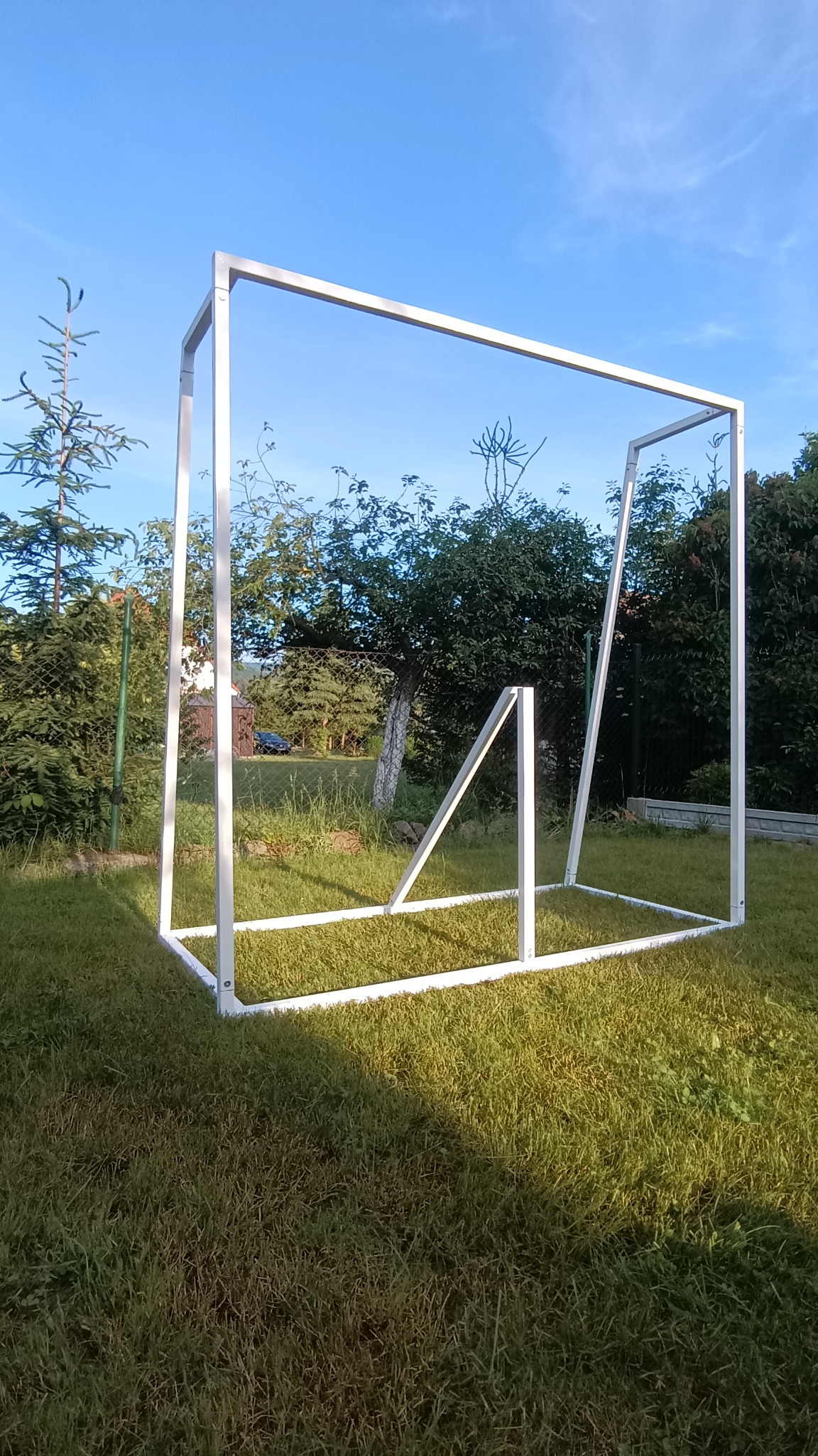 Metalowa konstrukcja w kształcie prostopadłościanu, prawdopodobnie początek pergoli, ustawiona na trawie w ogrodzie, w tle drzewa i błękitne niebo.