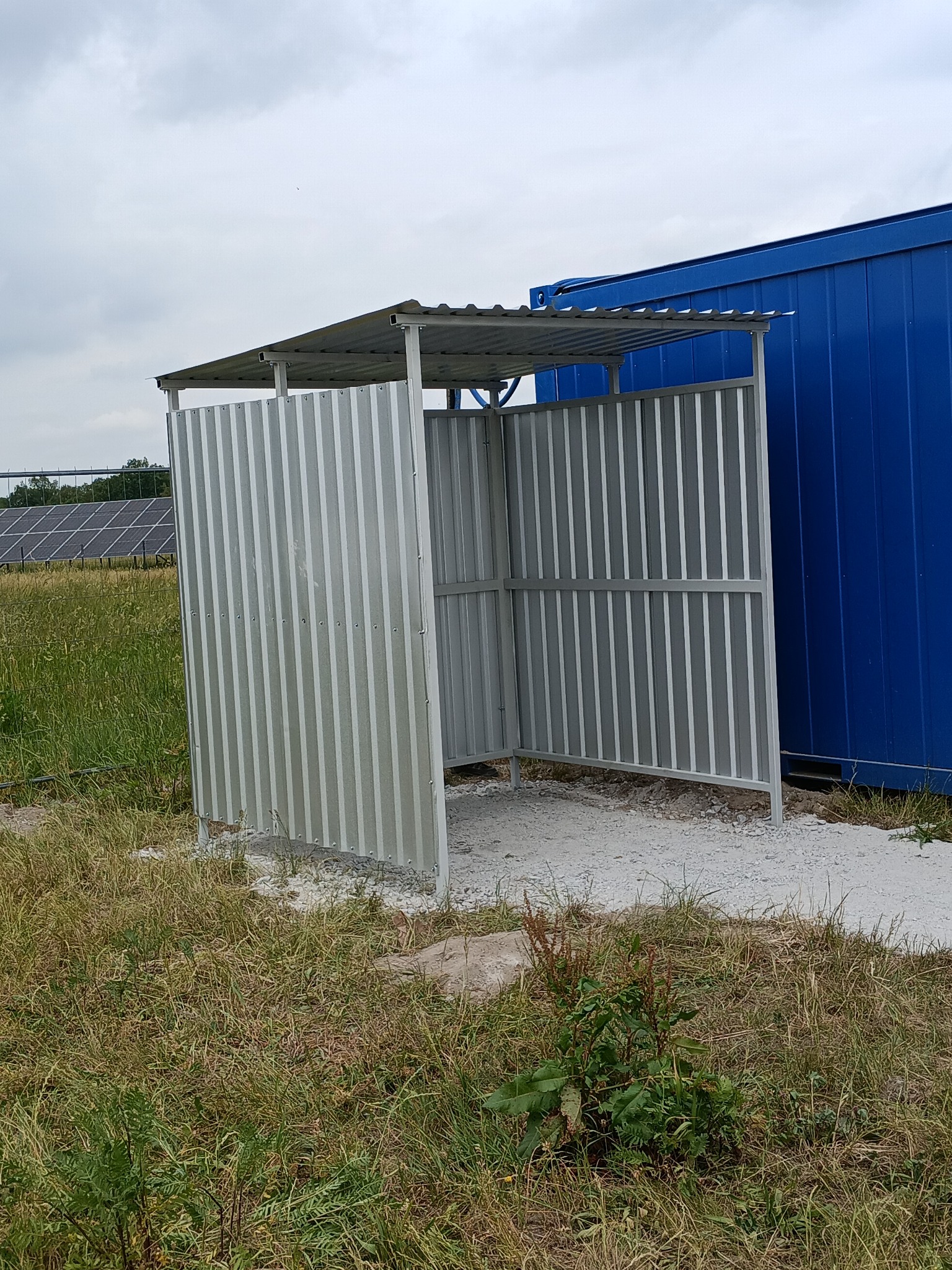 Metalowa konstrukcja osłonowa z zadaszeniem, wykonana z blachy trapezowej, stojąca na żwirowym podłożu obok niebieskiego kontenera, z widoczną trawą i w oddali panele słoneczne.