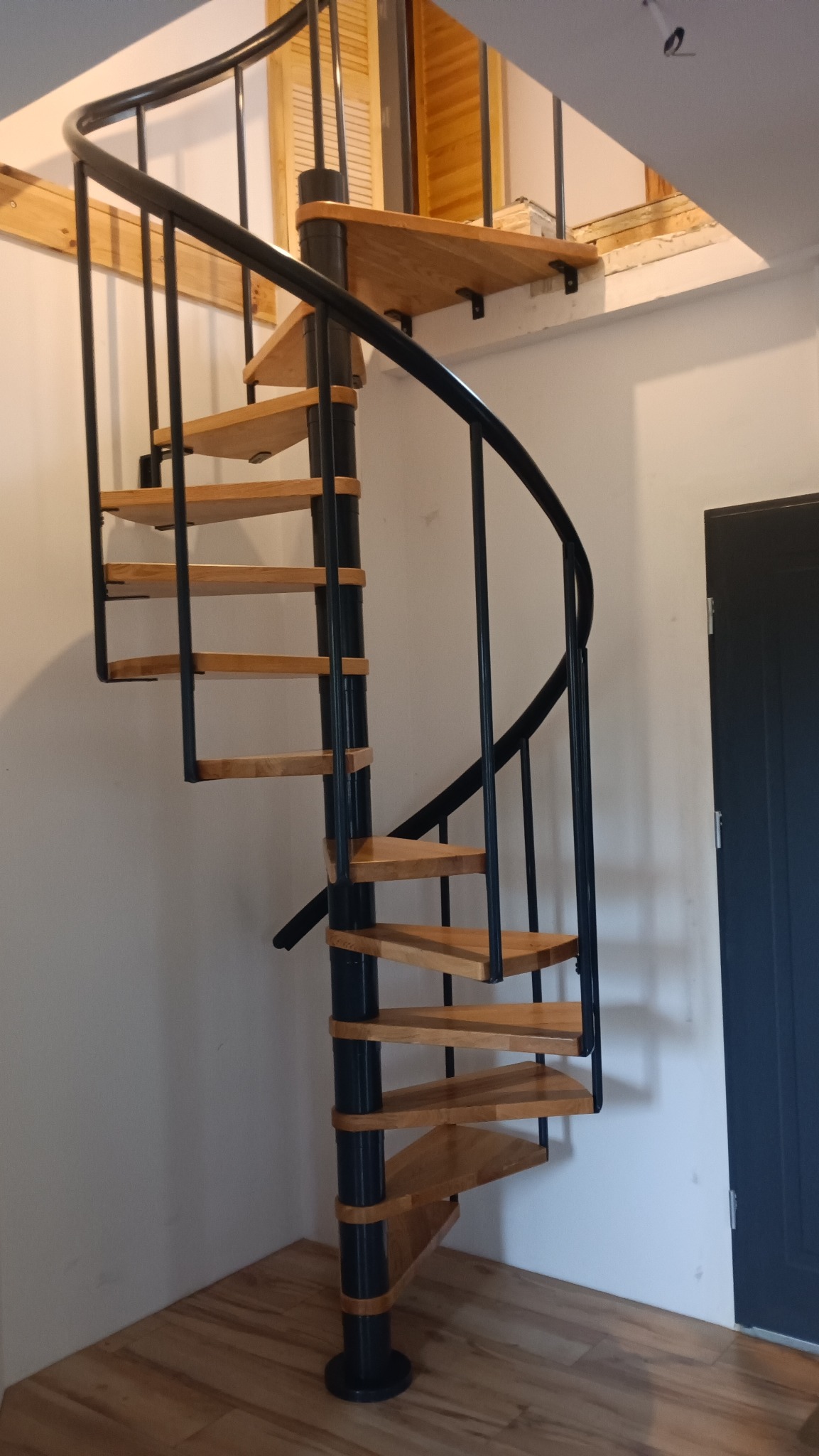 Spiralne schody z drewnianymi stopniami i czarną metalową konstrukcją w jasnym wnętrzu, widoczne drzwi i drewniane żaluzje w tle.