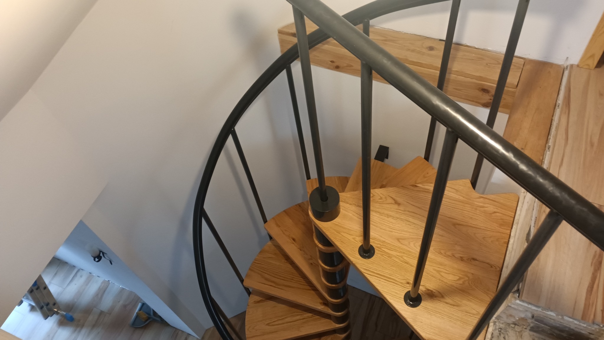 Widok z góry na drewniane schody spiralne z czarną metalową balustradą, w trakcie montażu, z widocznymi niedokończonymi elementami wykończenia podłogi i ścian.