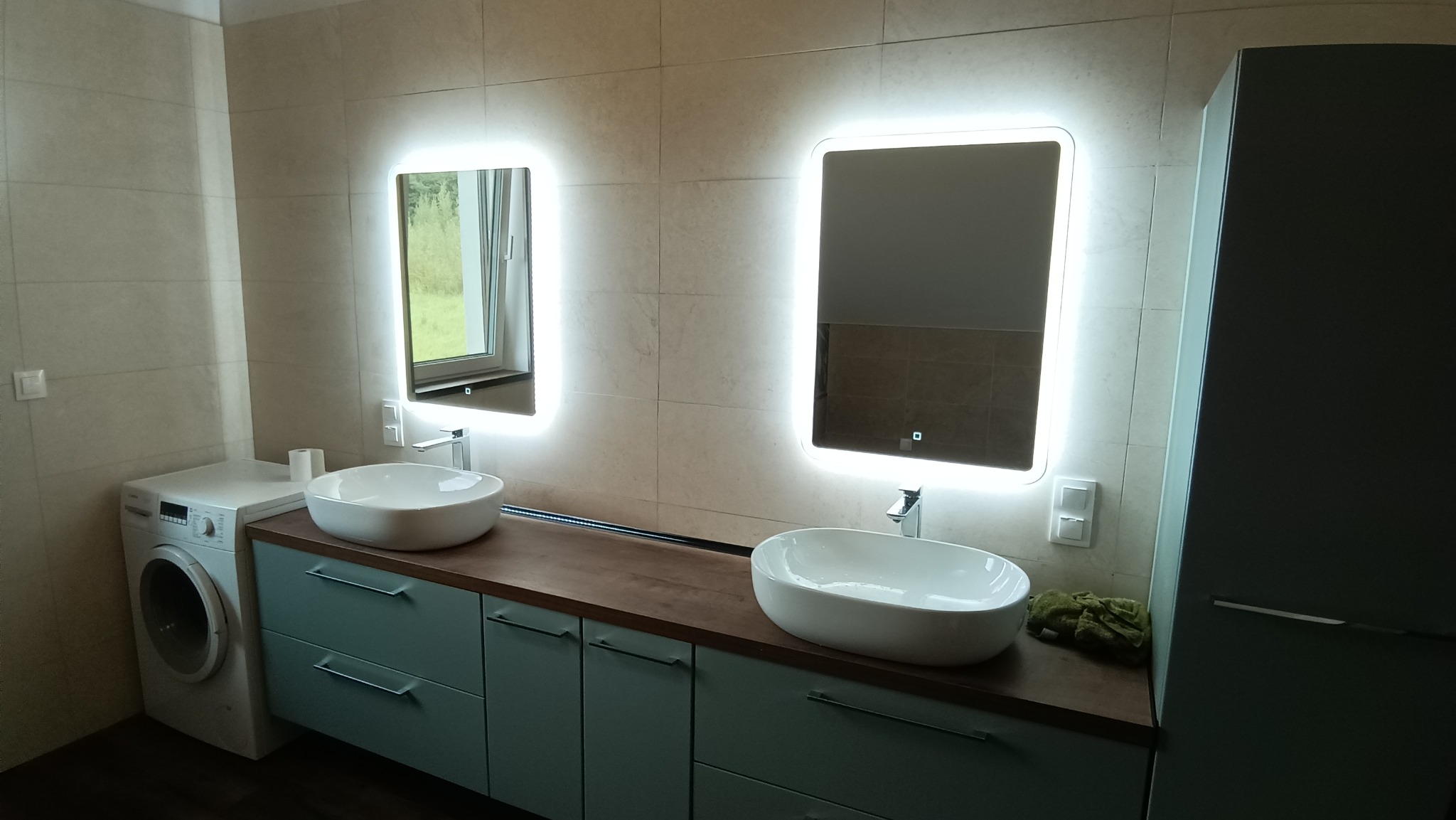 Nowoczesna łazienka z dwoma owalnymi umywalkami nablatowymi, podświetlanymi lustrami, szafkami w odcieniach błękitu i pralką.