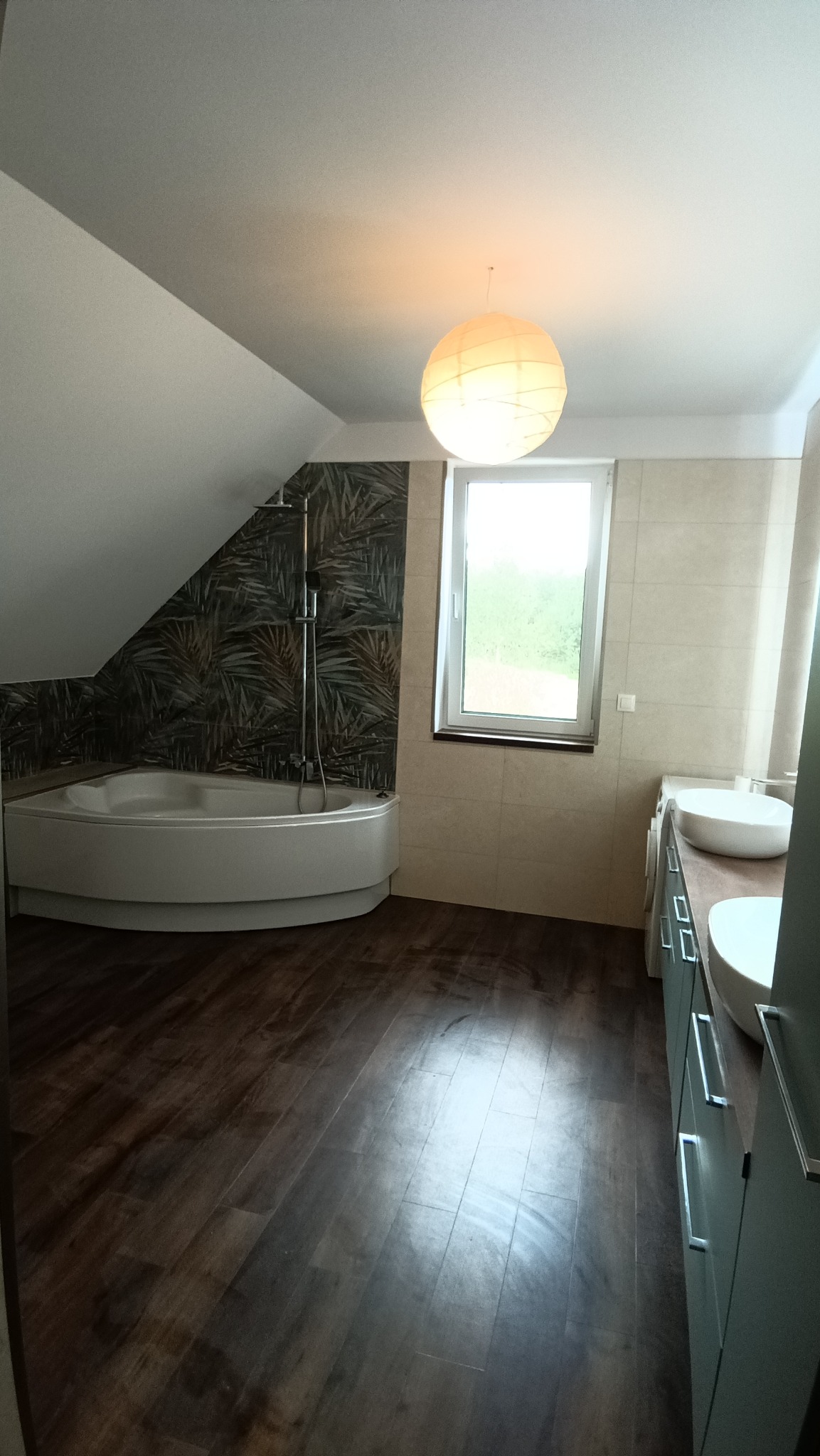 Nowoczesna łazienka na poddaszu z narożną wanną, deszczownicą, umywalkami nablatowymi i podświetlaną kulistą lampą.